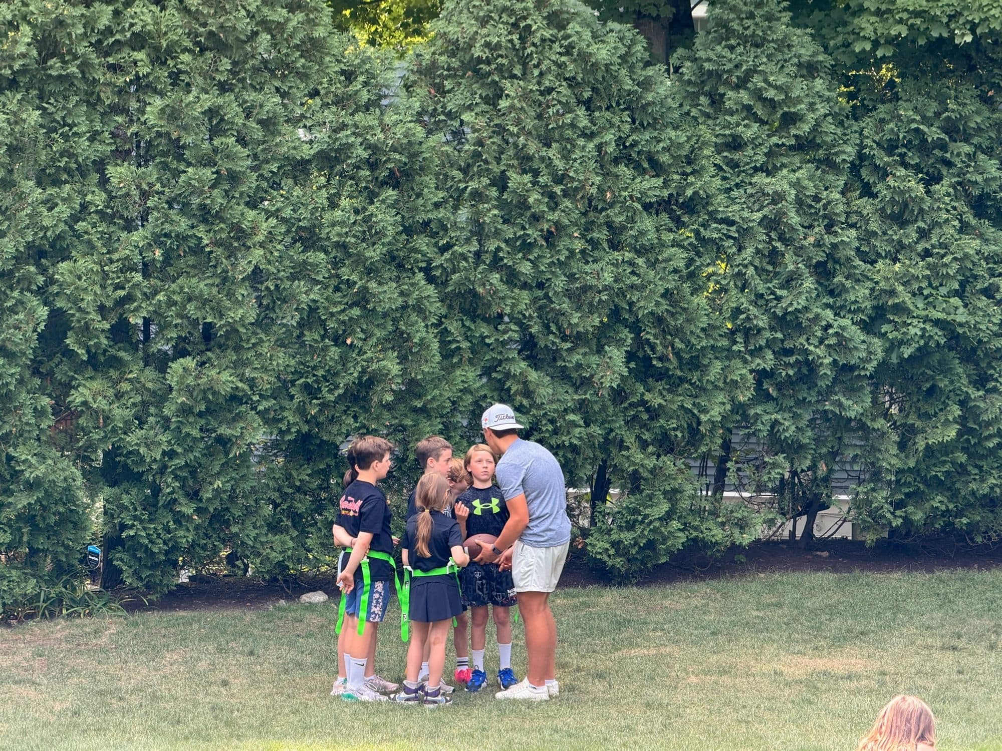 A coach crouched in a huddle with five campers wearing flag football belts at the edge of the field.