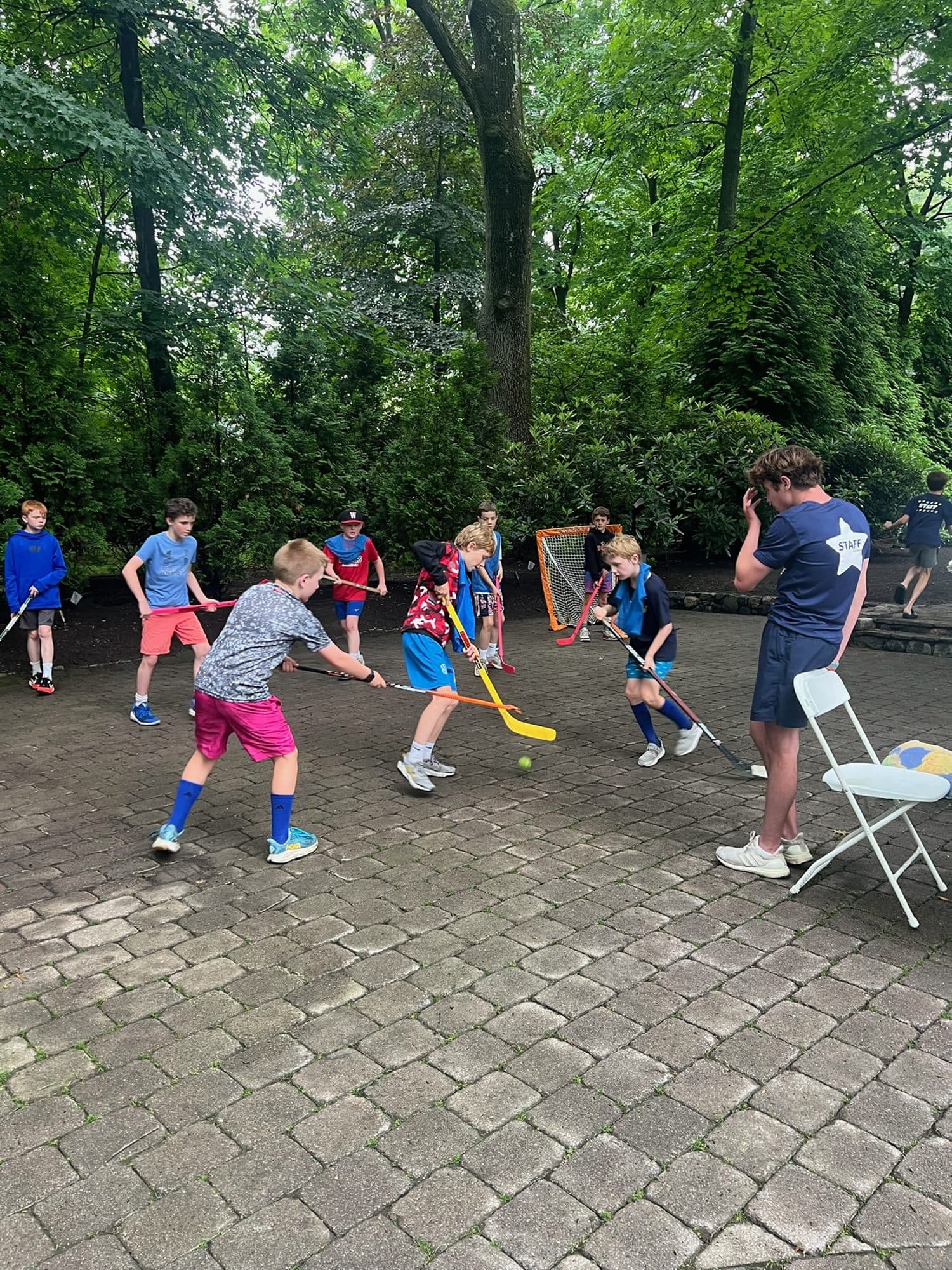 Campers playing floor hockey on a stone patio, sticks down around a yellow ball, with a coach watching from the sideline.