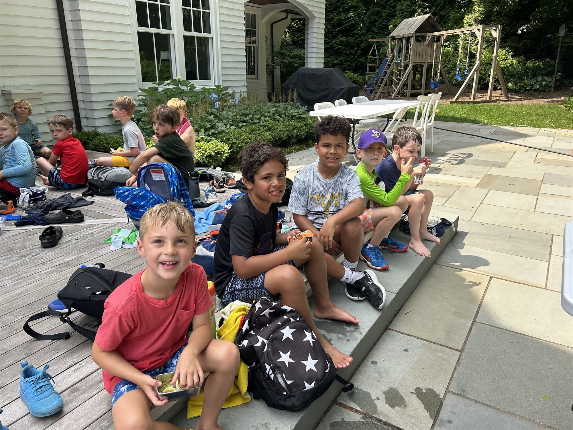 Campers sitting on a patio step with backpacks and snacks, smiling during a snack break.